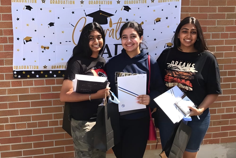 Drei Schülerinnen des West Humber Collegiate Institute stehen vor einer Abschlusskulisse und halten stolz ihre Diplome.