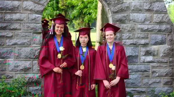 Drei Schülerinnen der West Nottingham Academy stehen in roten Abschlussroben vor einer Steinmauer mit grüner Vegetation.
