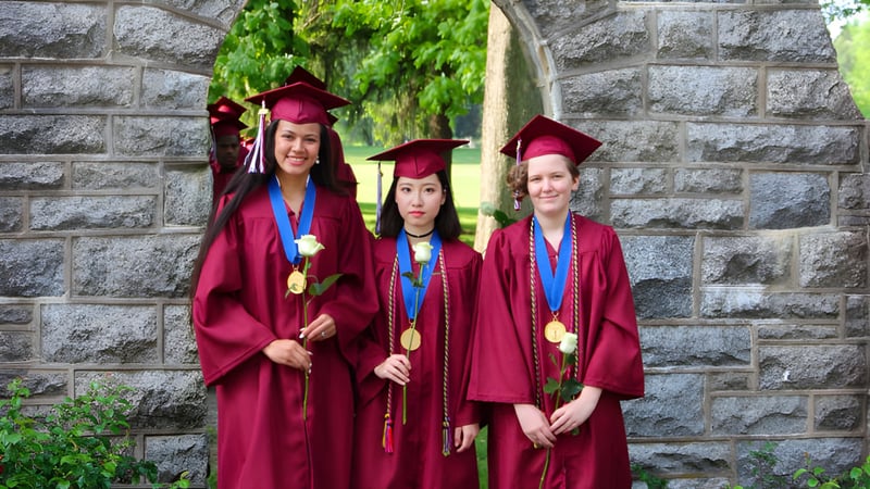 Drei Schülerinnen der West Nottingham Academy stehen in roten Abschlussroben vor einer Steinmauer mit grüner Vegetation.