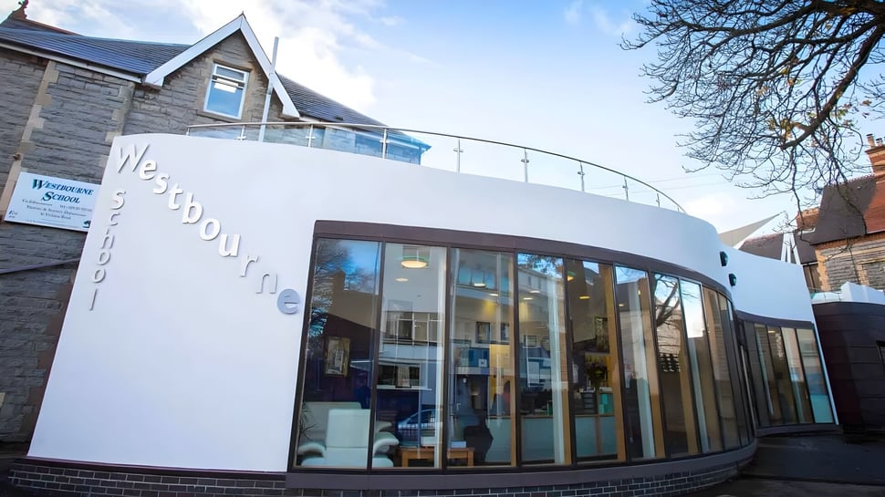 Das moderne Gebäude der Westbourne School mit einer Glasfassade steht vor Bäumen unter klarem Himmel.