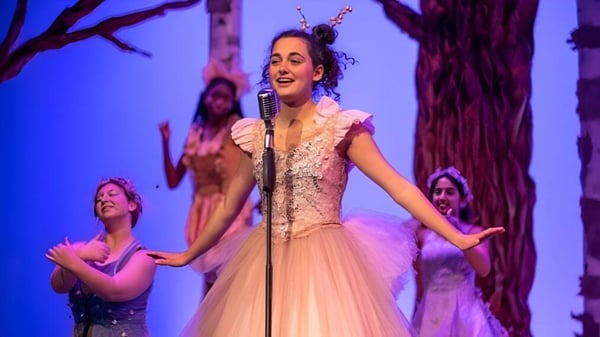 Eine Frau in einem mit Spitze verzierten Kleid singt auf der Bühne der Western Reserve Academy vor einem bunten Waldhintergrund mit Tänzern im Hintergrund.