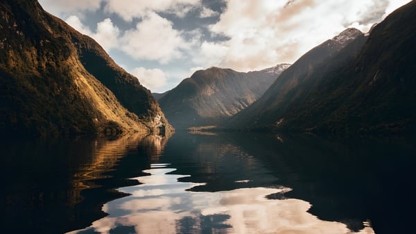 Ein ruhiger Fjord mit spiegelnder Wasseroberfläche und hohen Bergen im Hintergrund ist zu sehen.