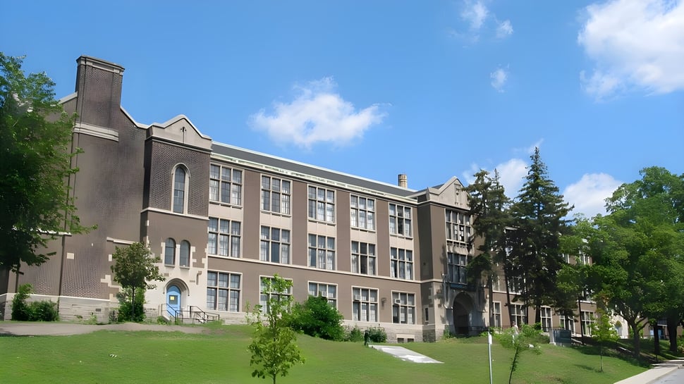 Ein mehrstöckiges Backsteingebäude der Western Technical-Commercial School steht umgeben von Bäumen und grasbewachsenem Vordergrund unter blauem Himmel.