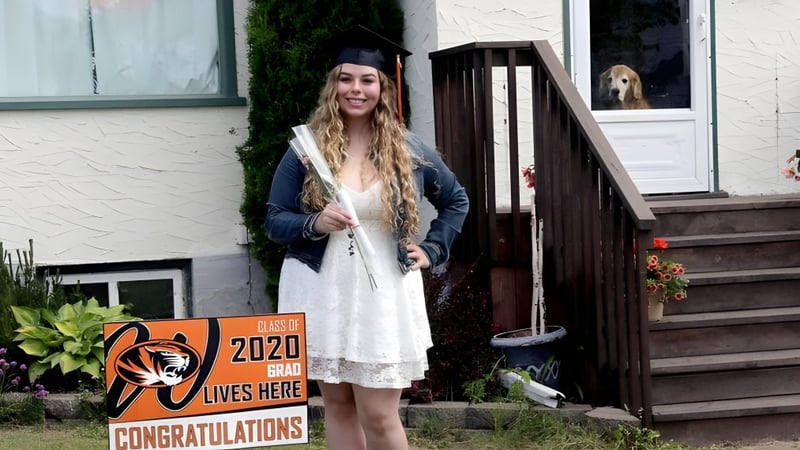 Eine Schülerin steht auf der Veranda eines rustikalen Holzgebäudes mit einem Schild, das '2020 lives here' und 'Congratulations' zeigt, auf dem Campus des Westgate Collegiate & Vocational Institute.