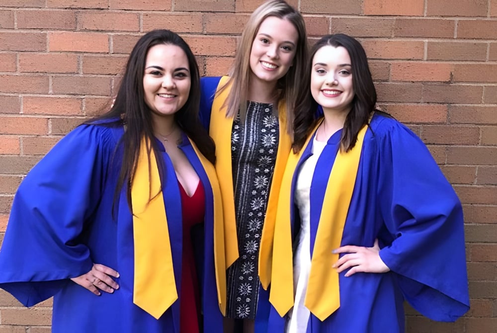 Drei Schülerinnen in blauen und gelben Abschlussroben posieren vor einer Backsteinwand auf dem Campus der Westisle Composite High School.