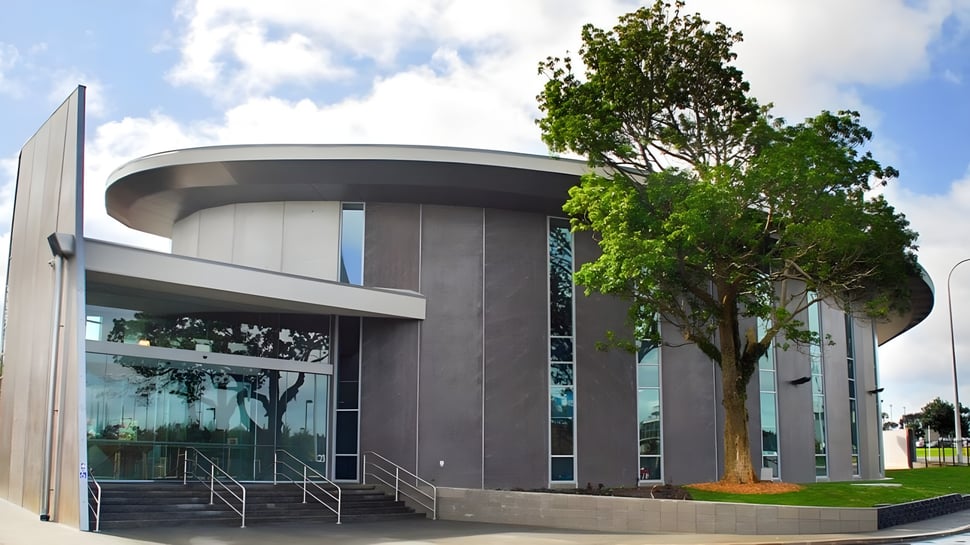 Das moderne Gebäude mit Glasfassade und großer Baum im Vordergrund auf dem Campus der Westlake Girls High School bei bewölktem Himmel.
