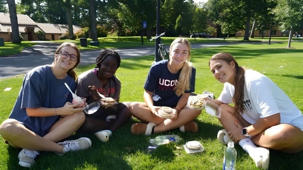 Vier Schülerinnen sitzen auf dem Gras im Park nahe der Westminster School mit Bäumen und Gebäuden im Hintergrund.