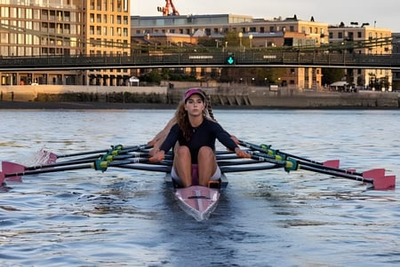 Eine Person rudert in einem pinken Boot auf dem Wasser vor der Kulisse der Westminster Secondary School bei Sonnenuntergang.