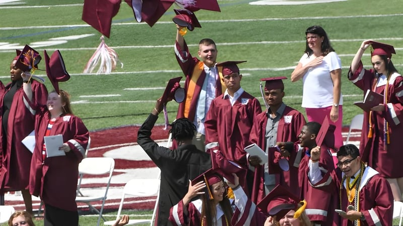 Absolventinnen und Absolventen der Westside Secondary School feiern auf einem Rasenfeld ihre Abschlussfeier in roten Talaren.