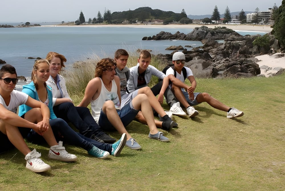 Eine Gruppe Schüler der Whakatane High School sitzt auf dem Gras nahe der felsigen Küste mit Wasser und Gebäuden im Hintergrund.