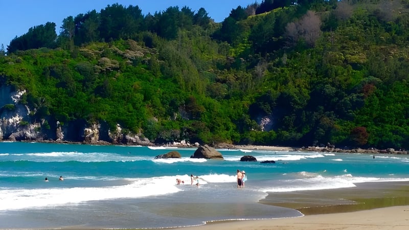 Menschen am Strand mit Blick auf das türkisblaue Meer und bewaldete Hügel am Küstengebiet nahe der Whangamata Area School.