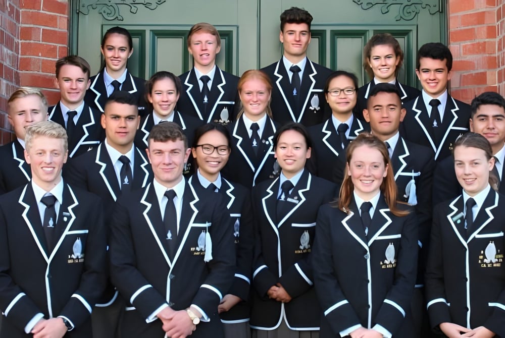 Eine Gruppe von Schülern der Whanganui Collegiate School steht in schwarzen Uniformen vor einem Backsteingebäude mit dekorativen Architekturdetails.
