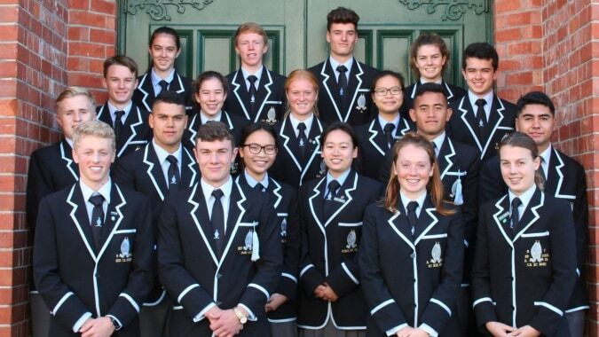 Eine Gruppe von Schülern der Whanganui Collegiate School steht in formeller Kleidung vor einem Backsteingebäude mit dekorativen Architekturmerkmalen.