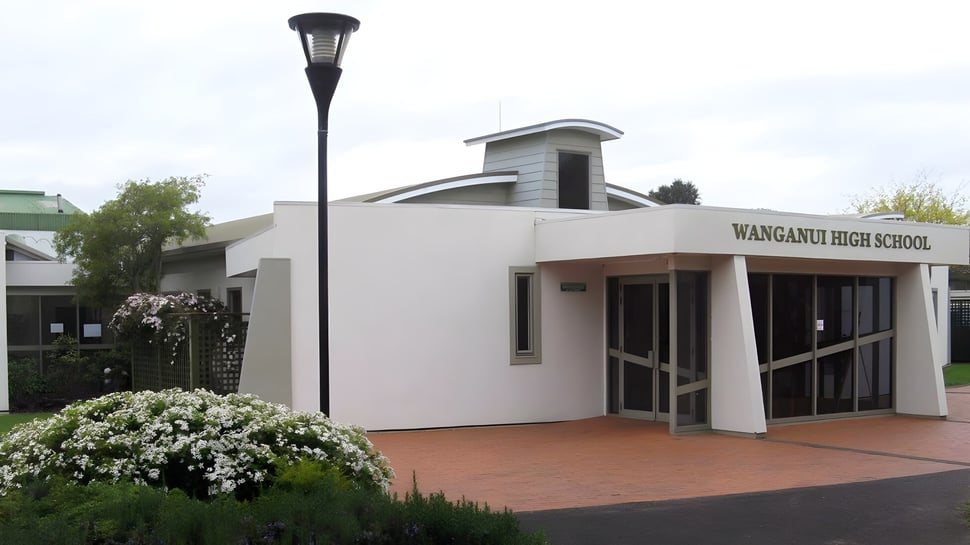 Das moderne weiße Schulgebäude der Whanganui High School mit dem Haupteingang und einer hohen Laterne im Vordergrund, umgeben von Grün und Blumen.
