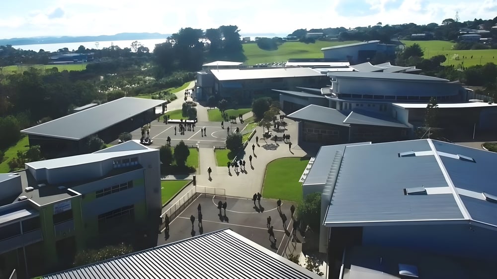 Der Campus von Whangaparāoa College mit modernen Gebäuden, Wegen und Grünflächen vor einer hügeligen Landschaft und einem Gewässer.
