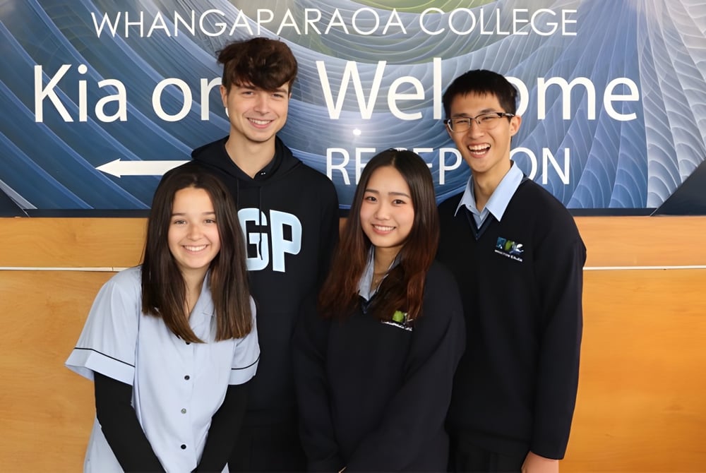 Eine Gruppe von vier Schülerinnen und Schülern posiert vor einem Banner mit 'Kia ora Welcome' auf dem Campus des Whangaparāoa College.