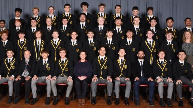 Eine große Gruppe von Schülern der Whangarei Boys’ High School sitzt in formeller Kleidung vor grauem Hintergrund.