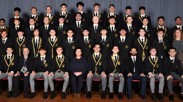 Eine große Gruppe von Schülern in formeller akademischer Kleidung sitzt in Reihen vor einem grauen Hintergrund auf dem Campus der Whangarei Boys’ High School.