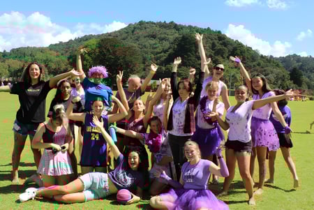 Eine Gruppe Schülerinnen der Whangarei Girls High School steht auf einem grasbewachsenen Feld vor grünen Hügeln unter blauem Himmel.