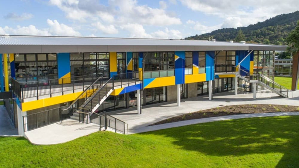 Das moderne und farbenfrohe Gebäude der Whangarei Girls High School steht auf einer Wiese mit grünen Hügeln und Bergen im Hintergrund.