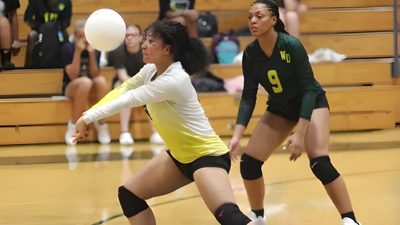 Zwei weibliche Volleyballspielerinnen der White Oaks Secondary School spielen auf dem Volleyballfeld mit Holzbänken im Hintergrund.