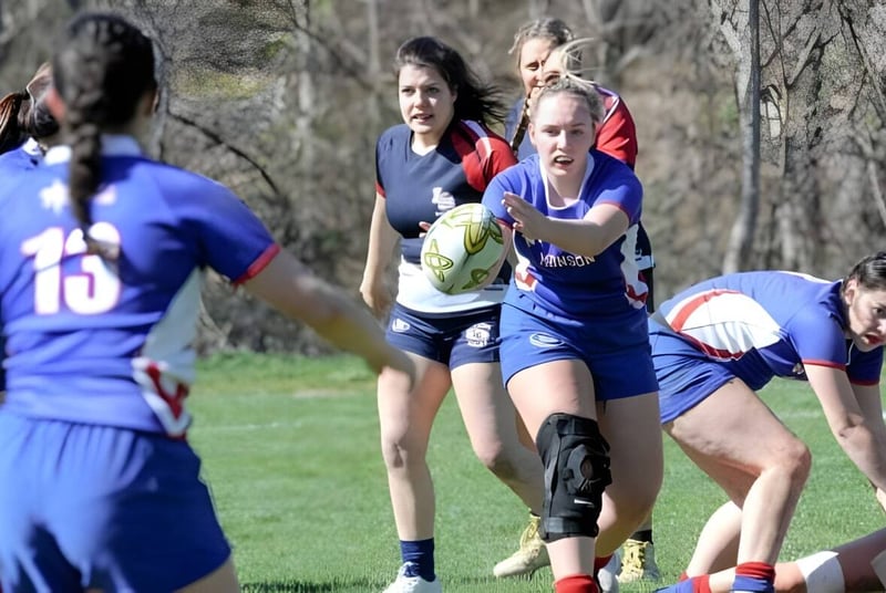 Weibliche Rugby-Spielerinnen der Wilbraham & Monson Academy beim Spiel auf einem Grasfeld mit Bäumen im Hintergrund.