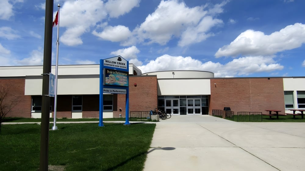 Das Backsteingebäude der Willow Creek Composite High School mit einem blauen Schild und kanadischer Flagge unter bewölktem Himmel.