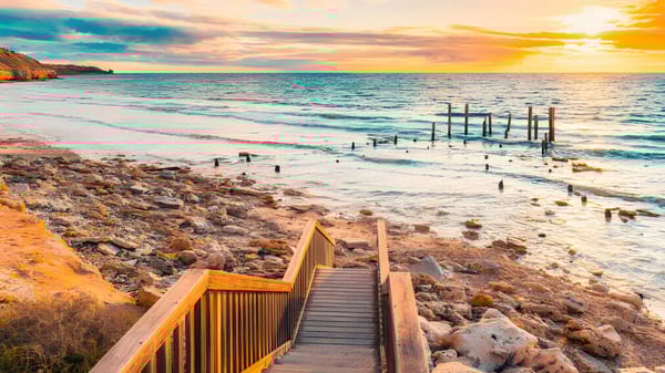Ein hölzerner Steg führt an einen felsigen Strand mit ruhigem Wasser unter einem farbenfrohen Sonnenuntergang bei Willunga High School.