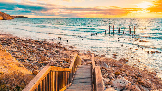 Ein hölzerner Weg führt zum felsigen Strand mit ruhigem türkisem Wasser und einem Sonnenuntergangshimmel, der bei Willunga High School zu sehen ist.