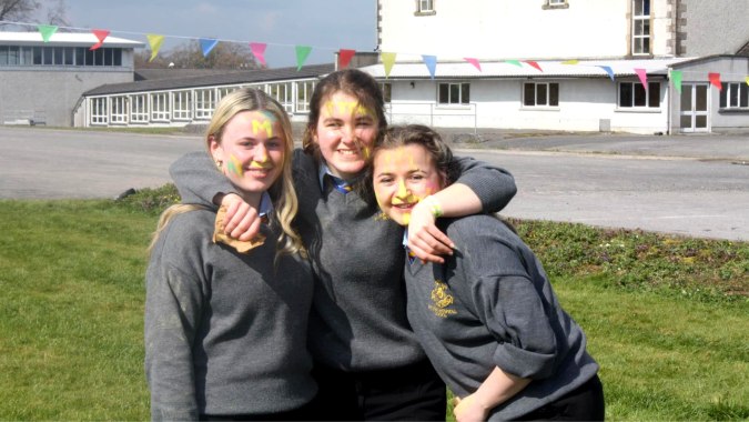 Drei Schülerinnen der Wilson's Hospital School stehen zusammen und umarmen sich vor einer farbenfrohen Fahnenwand.