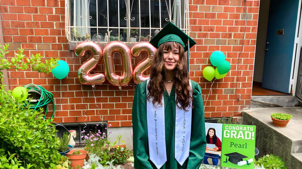 Eine junge Frau in grünem Abschlussmantel steht vor einer Backsteinwand mit Ballons, die 2022 formen, auf dem Campus der Winchendon School in New York City.