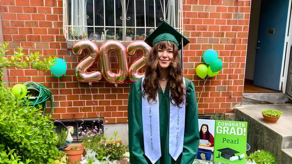 Eine junge Frau in grünem Abschlussmantel steht vor einer Backsteinwand mit Ballons, die 2022 formen, auf dem Campus der Winchendon School in New York City.