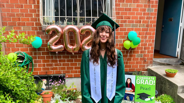 Eine Schülerin der Winchendon School in New York City steht in grünem Abschlussmantel vor einer Ziegelwand mit bunten Luftballons und einem Gratulationsschild.