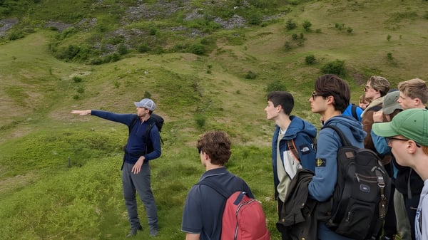 Eine Gruppe Schüler steht auf einem grasbewachsenen Hügel mit üppiger Vegetation vor einer landschaftlichen Kulisse auf dem Campus von Winchester College.
