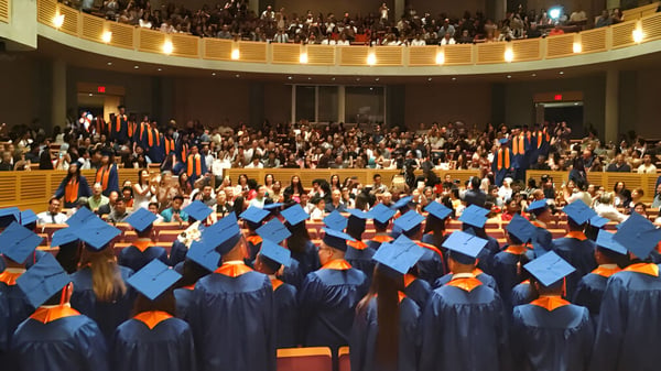 Absolventinnen und Absolventen der Windermere Secondary School sitzen in einer feierlich dekorierten Aula in blauen und orangen Abschlusskleidern.