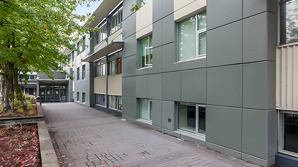 Das mehrstöckige Gebäude der Windsor Secondary School mit grauer Fassade steht an einem gepflasterten Weg und ist von Bäumen umgeben.