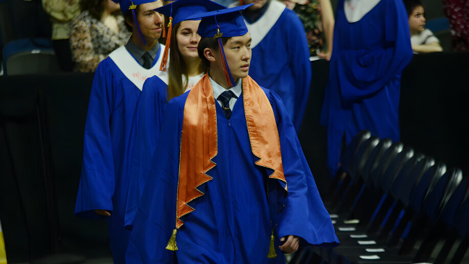 Eine Gruppe von Absolventinnen und Absolventen in blauen und orangen Abschlussroben steht vor der Winston Churchill High School.