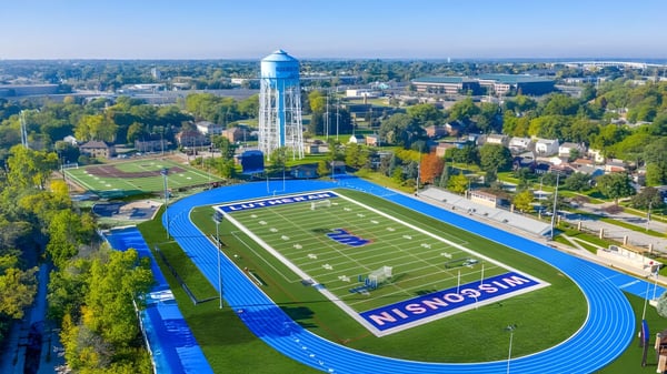 Das große Footballfeld mit blauer Laufbahn auf dem Gelände der Wisconsin Lutheran High School zeigt im Hintergrund die Stadtsilhouette mit einem hohen Wasserturm.