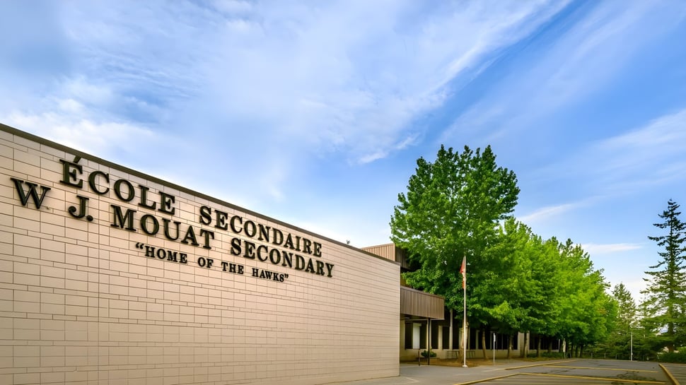 Das große Gebäude der W.J. Mouat Secondary School ist von grünen Bäumen umgeben und steht unter einem blauen Himmel mit Wolken.