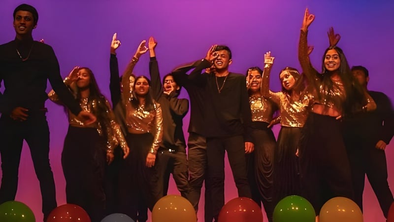 Schüler des Woburn Collegiate Institute stehen auf der Bühne in gold-schwarzen Kostümen mit erhobenen Armen vor lila Hintergrund.