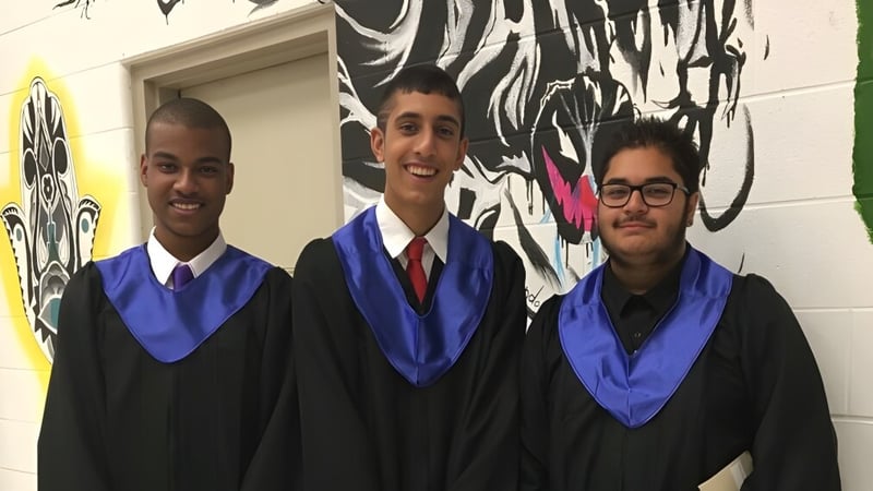 Drei Absolventinnen und Absolventen in Talaren stehen vor einem bunten Wandbild auf dem Campus des Woodbridge College.