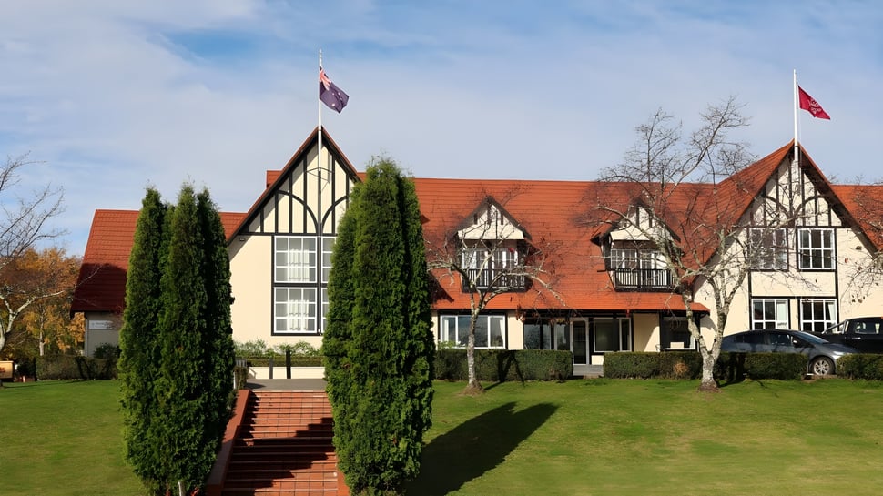 Das Tudor-Gebäude der Woodford House mit roten Ziegeldächern und Fachwerkfassaden steht auf gepflegtem Rasen umgeben von Zypressen.