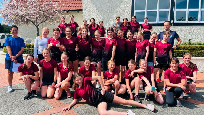 Eine Gruppe Schülerinnen in roten Uniformen posiert vor einem Gebäude mit blühendem Baum auf dem Campus der Woodford House.