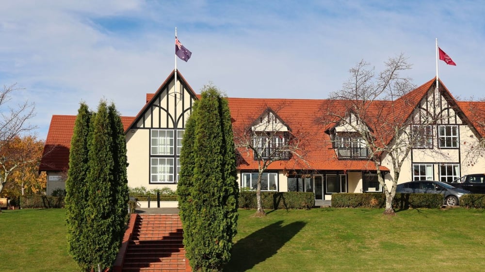 Das Tudor-Gebäude von Woodford House steht auf einem gepflegten Rasen, umgeben von Zypressen und einem gepflasterten Weg zum Eingang.