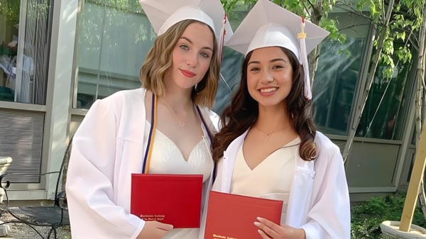 Zwei Schülerinnen der Woodlands Academy of the Sacred Heart in Abschlusskleidung halten stolz ihre Diplome vor einem Gebäude mit Grünfläche.