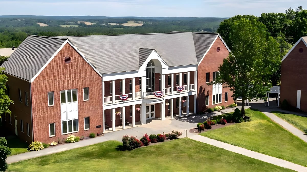 Das mehrstöckige Backsteingebäude der Woodstock Academy steht vor grünen Hügeln und umgeben von üppiger Vegetation.