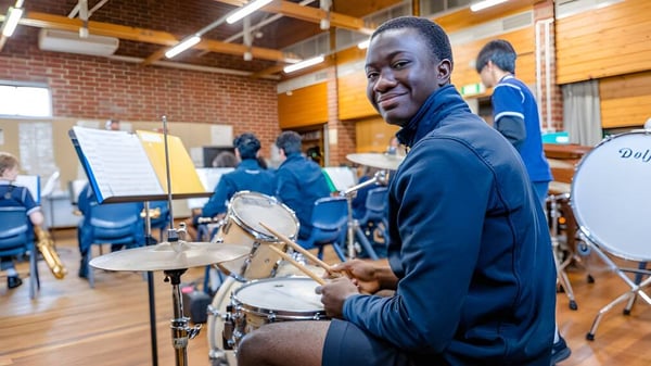 Ein Mann trägt eine blaue Jacke und spielt Schlagzeug im Musikstudio der Woodville High School mit weiteren Musikern im Hintergrund.