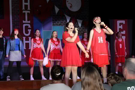 Eine Gruppe von Schülern der Woolgoolga High School tritt in roten Cheerleader-Kostümen auf einer Bühne mit mehreren Werbebannern im Hintergrund auf.