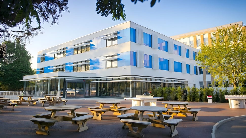 Das moderne mehrstöckige Gebäude mit Glasfassade der Worthgate School Canterbury ist von einem Außenbereich mit Holztischen umgeben.