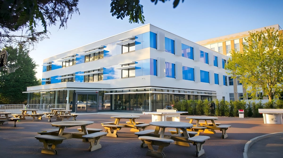 Das moderne mehrstöckige Gebäude mit Glasfassade der Worthgate School Canterbury ist von einem Außenbereich mit Holztischen umgeben.